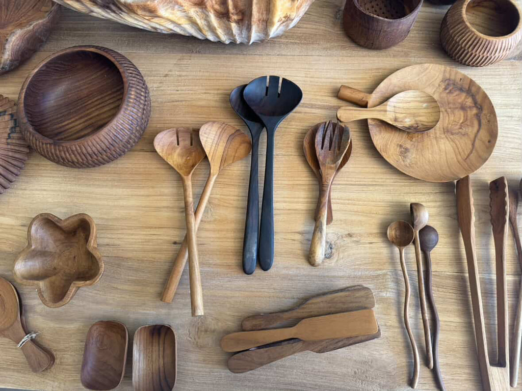 A collection of handcrafted wooden serving utensils and bowls arranged on a light wood tabletop, including spoons, forks, chopsticks, a serving tray, and small bowls.