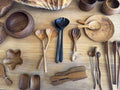 A collection of handcrafted wooden serving utensils and bowls arranged on a light wood tabletop, including spoons, forks, chopsticks, a serving tray, and small bowls.
