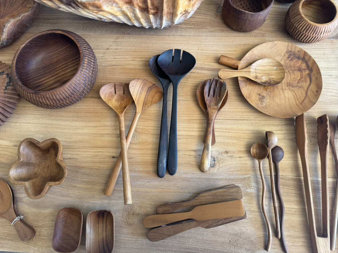 A collection of handcrafted wooden serving utensils and bowls arranged on a light wood tabletop, including spoons, forks, chopsticks, a serving tray, and small bowls.