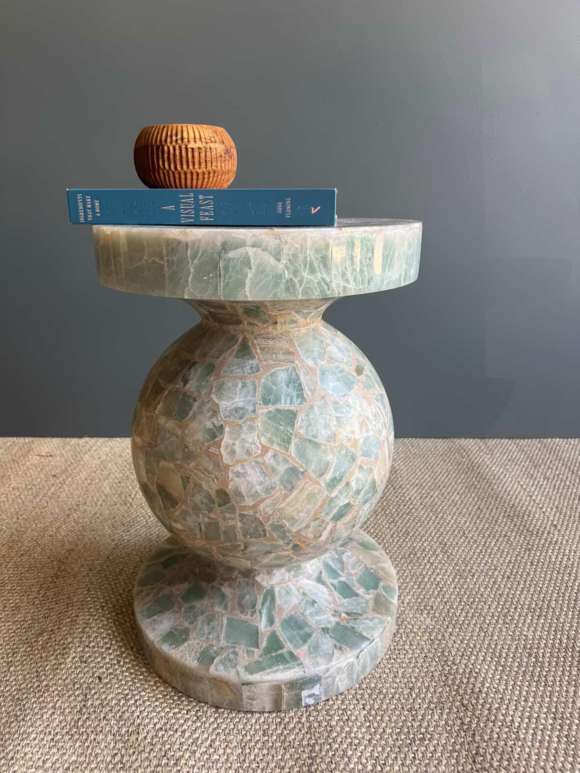 HC Homewares round marble side table with a mosaic green and beige stone pattern, styled with a blue book and a small wooden bowl on top.