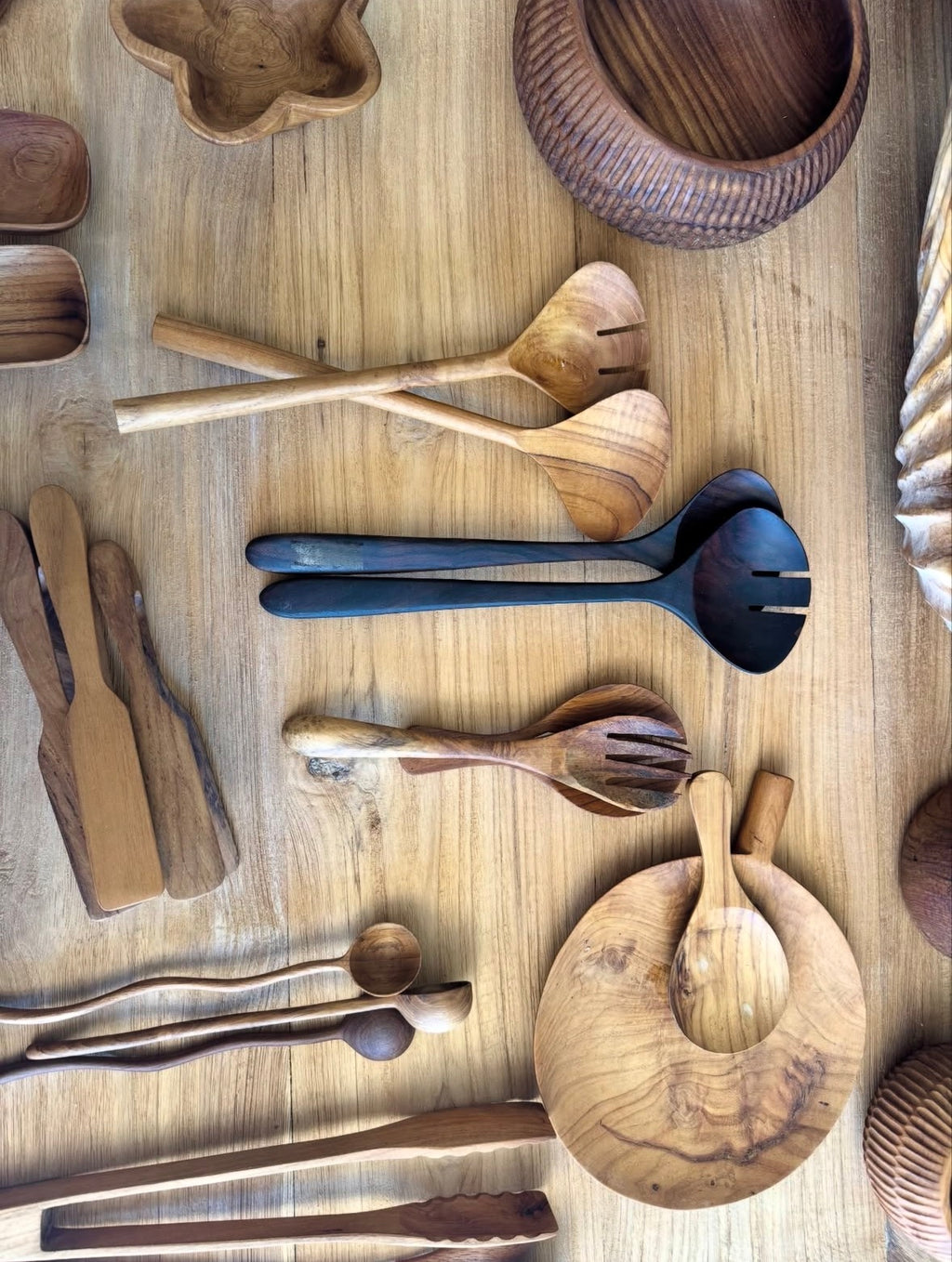 A collection of handcrafted wooden kitchen utensils, including serving spoons, salad servers, tongs, and a round wooden board, arranged on a light wood tabletop.